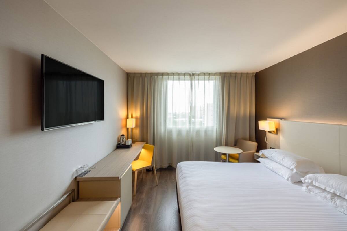 Une chambre d’hôtel comprenant un lit, un bureau, une chaise, une petite table, un téléviseur fixé au mur et une fenêtre avec des rideaux.