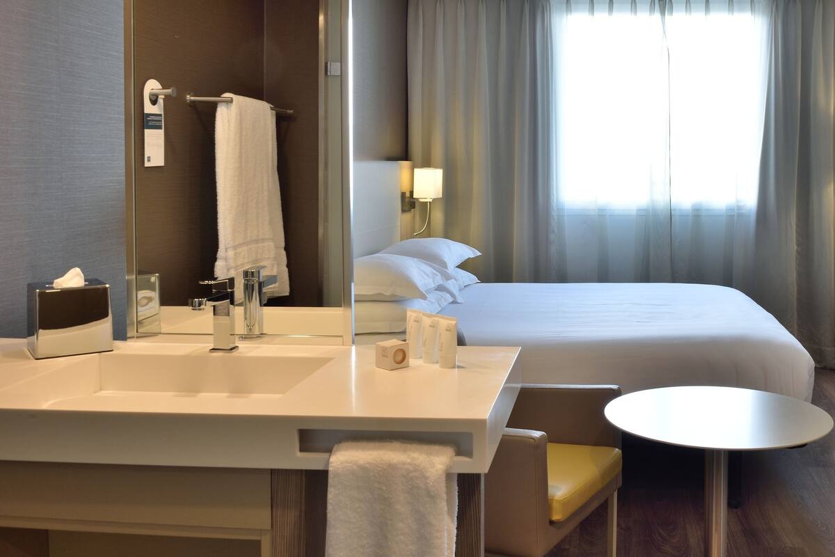 Une chambre d’hôtel comprenant un lit, une table de chevet, une chaise, un bureau, une lampe, une télévision et une salle de bain avec un lavabo et un porte-serviettes.