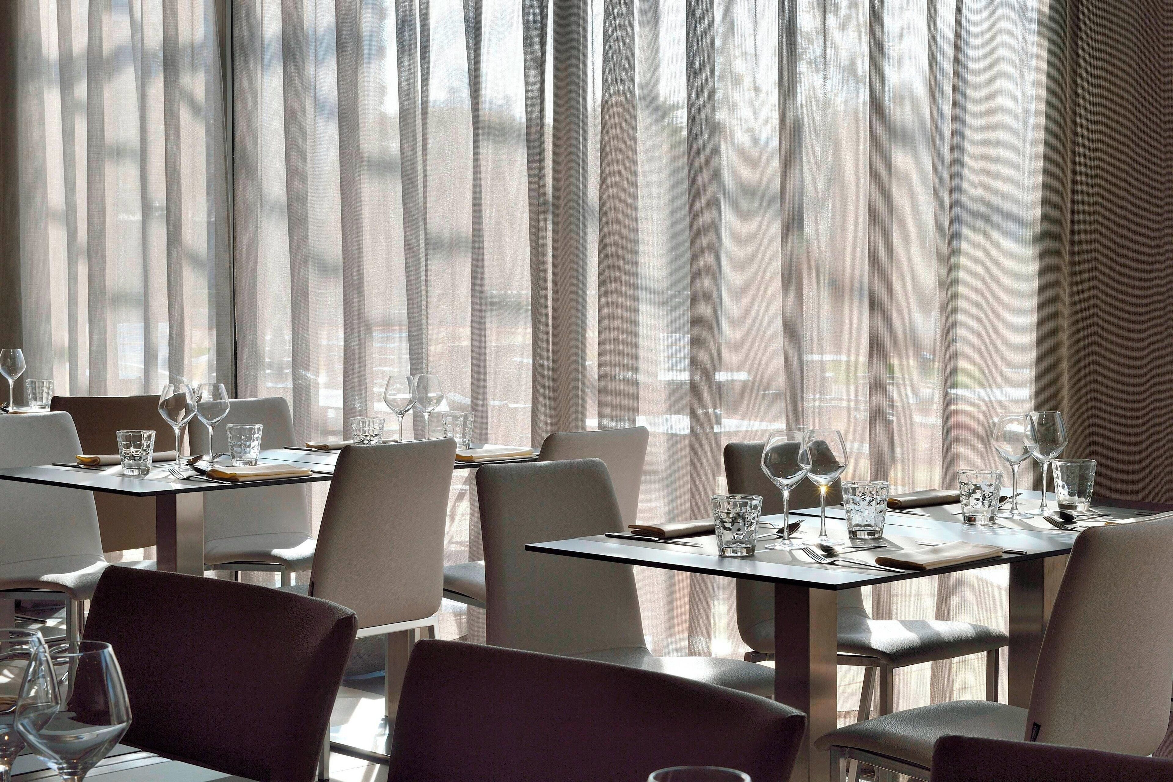 Un restaurant avec des tables dressées pour le service, de grandes fenêtres ornées de rideaux légers et une vue sur la rue.