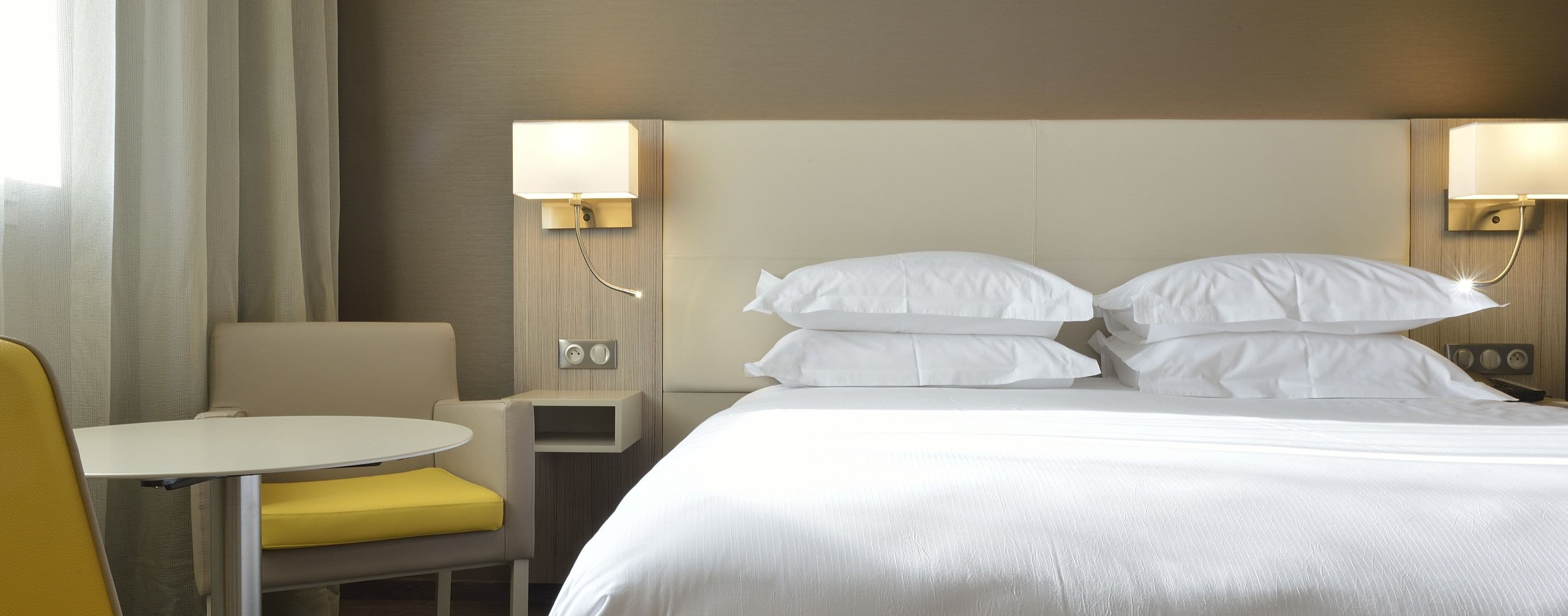 Una habitación de hotel con cama, dos lámparas de noche, una mesita con una silla y un cojín amarillo.