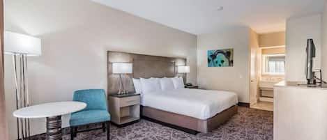 A hotel room with a bed, a desk, a chair, a small table, and a painting on the wall.