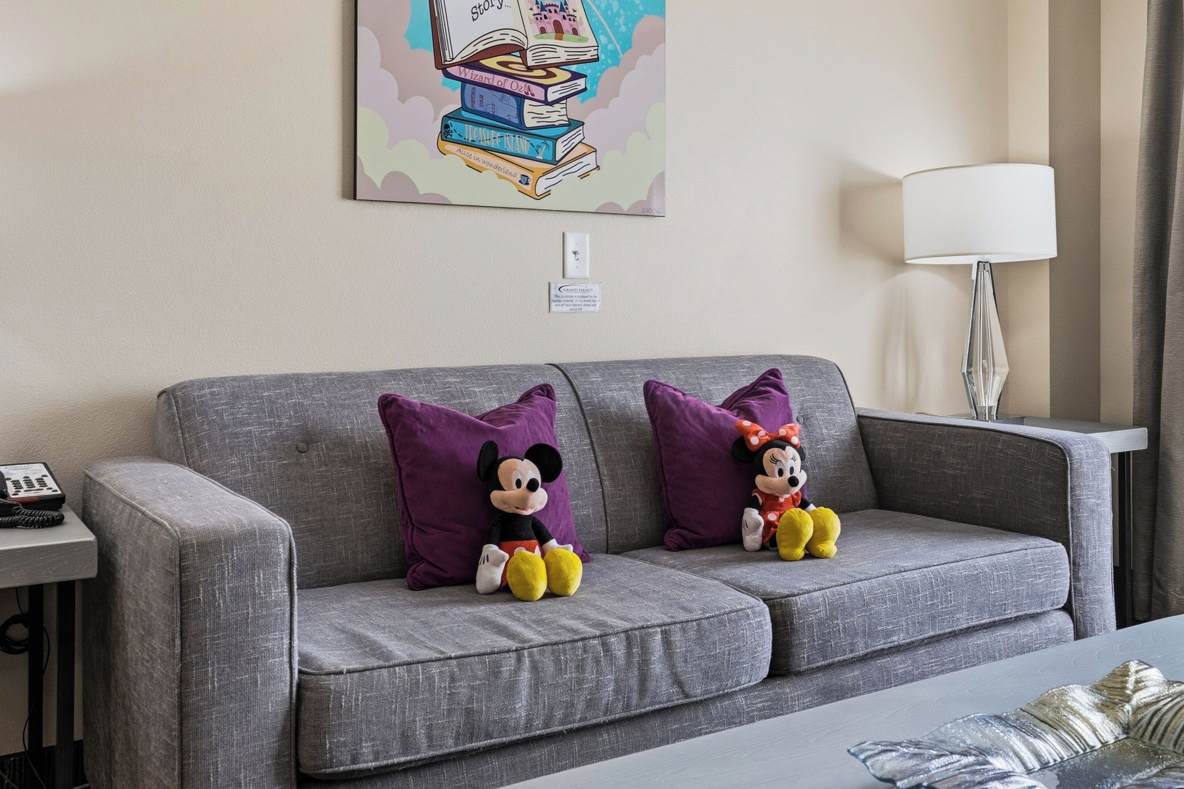 A grey sofa with two purple pillows, two plush toys, a lamp, and a framed picture on the wall.