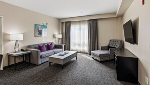 A hotel room with a grey sofa, a coffee table, a patterned armchair, a flat-screen TV, and a window with curtains.