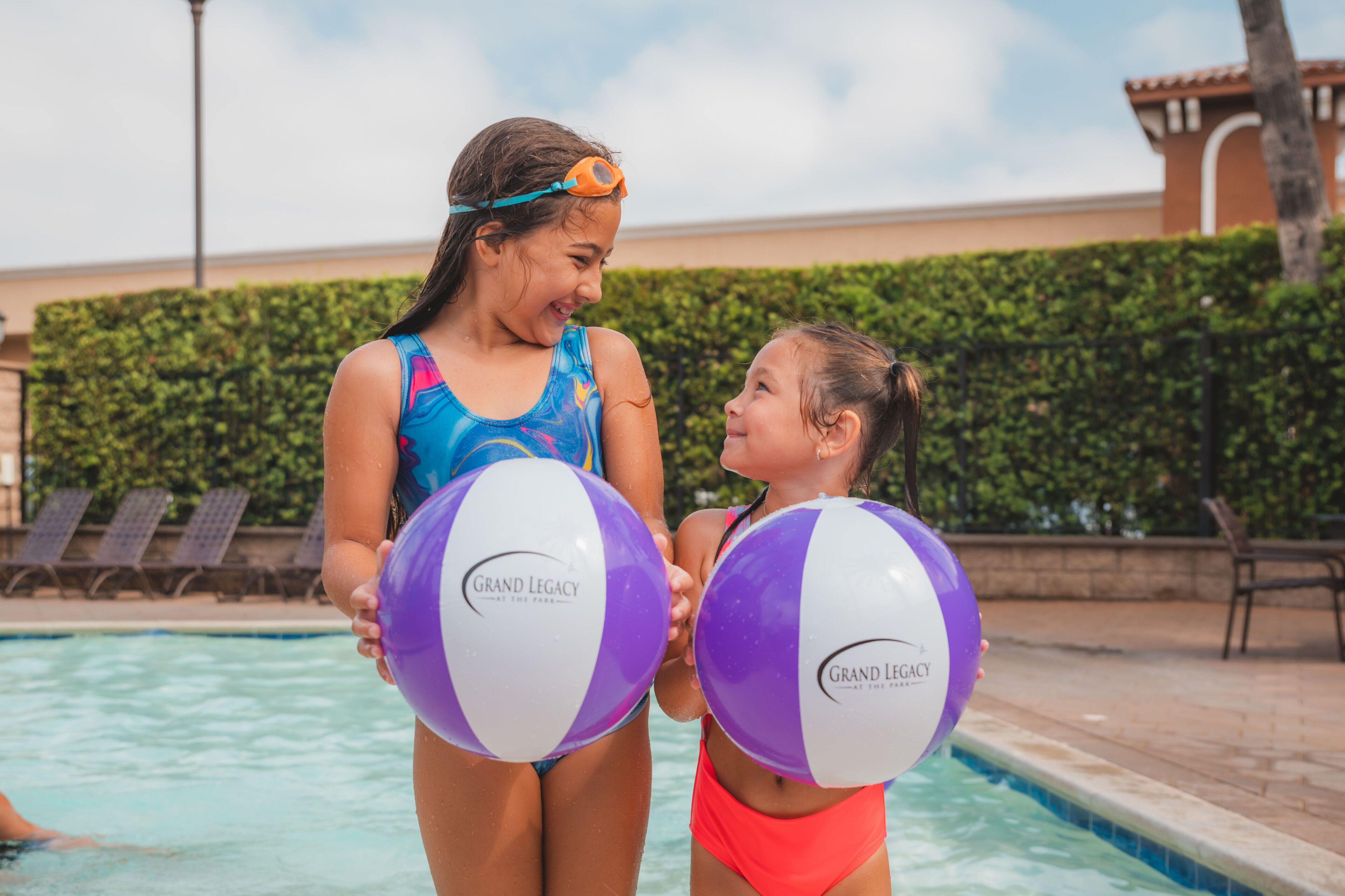 Two people holding beach balls by a pool with a hedge and chairs in the background.