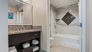 Un baño moderno con encimera de granito, mueble de tocador de madera oscura, cabina de ducha con mampara de vidrio y pared de ducha con azulejos de mármol.