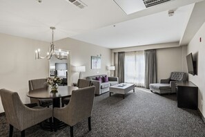 A hotel room with a dining table, chairs, a sofa, a coffee table, a TV, and a large window with curtains. - Grand Legacy At the Park (Anaheim)