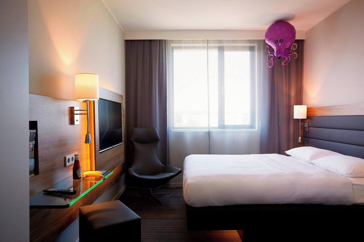 Habitación de hotel con cama, televisor, silla, escritorio con lámpara y ventana con cortinas.