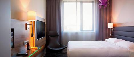 A hotel room with a bed, a TV, a chair, a desk with a lamp, and a window with curtains.