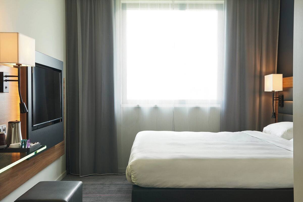 Habitación de hotel con cama, televisor, escritorio con lámpara y ventana con cortinas.