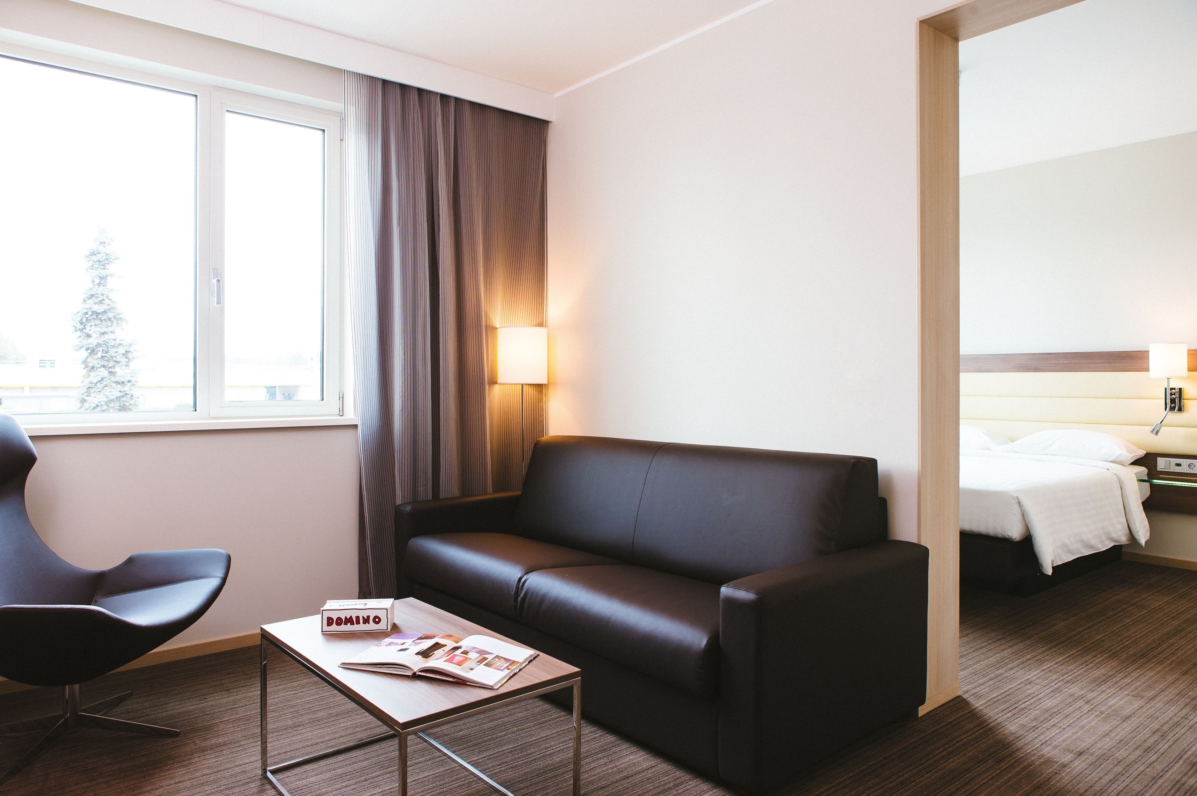 Une chambre d’hôtel avec un canapé en cuir, une petite table sur laquelle repose un magazine, une chaise, une fenêtre avec des rideaux et un lit doté d’une tête de lit blanche.