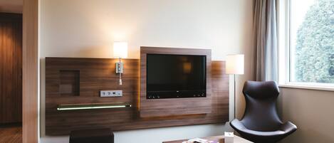 A modern hotel room with a flat-screen TV mounted on a wooden panel, a comfortable armchair, a small table with a magazine, and a window with curtains.