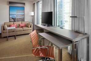 A modern hotel room with a sofa, a desk with a flat-screen TV, a chair, and a large window with a view of a cityscape.