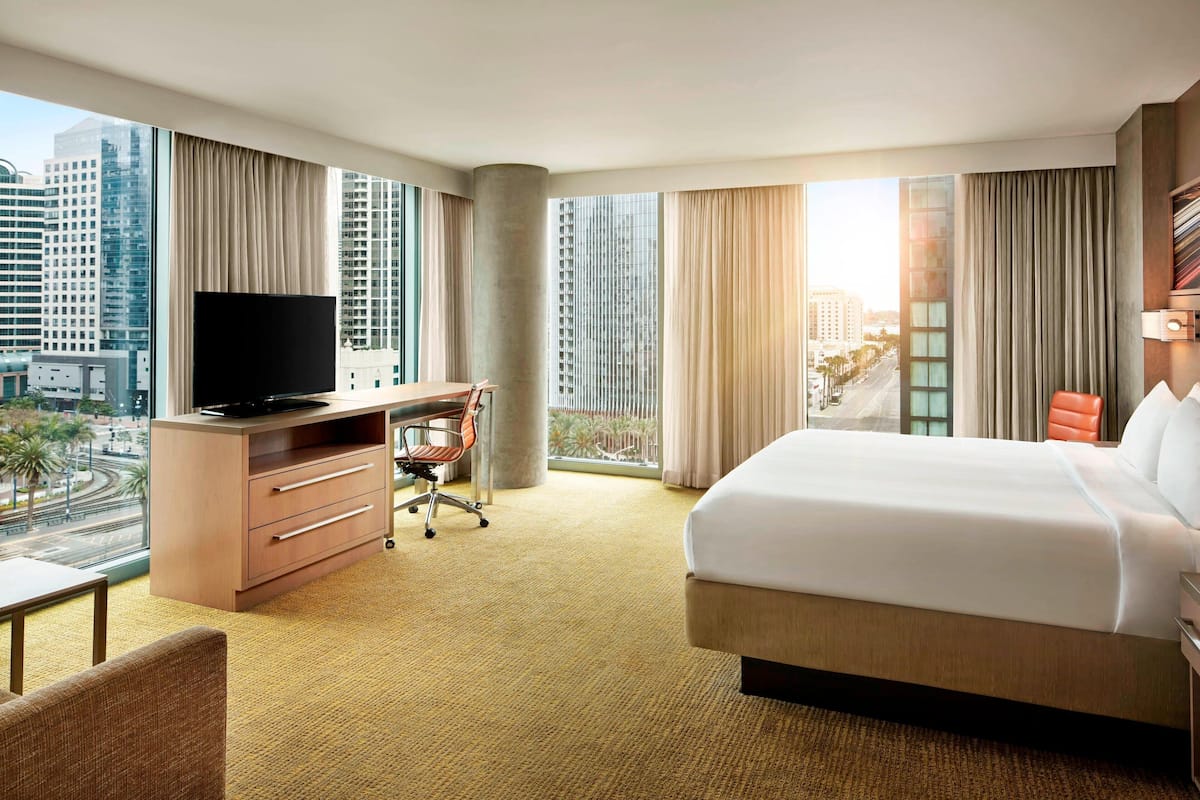 Habitación de hotel con una cama grande, un escritorio con televisor, una silla y vistas a un paisaje urbano a través de amplias ventanas.