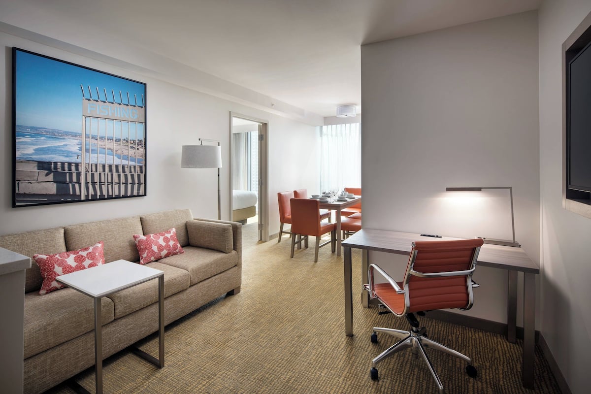 Habitación de hotel con sofá, escritorio, silla, mesa de comedor y un cuadro grande con una escena de playa.