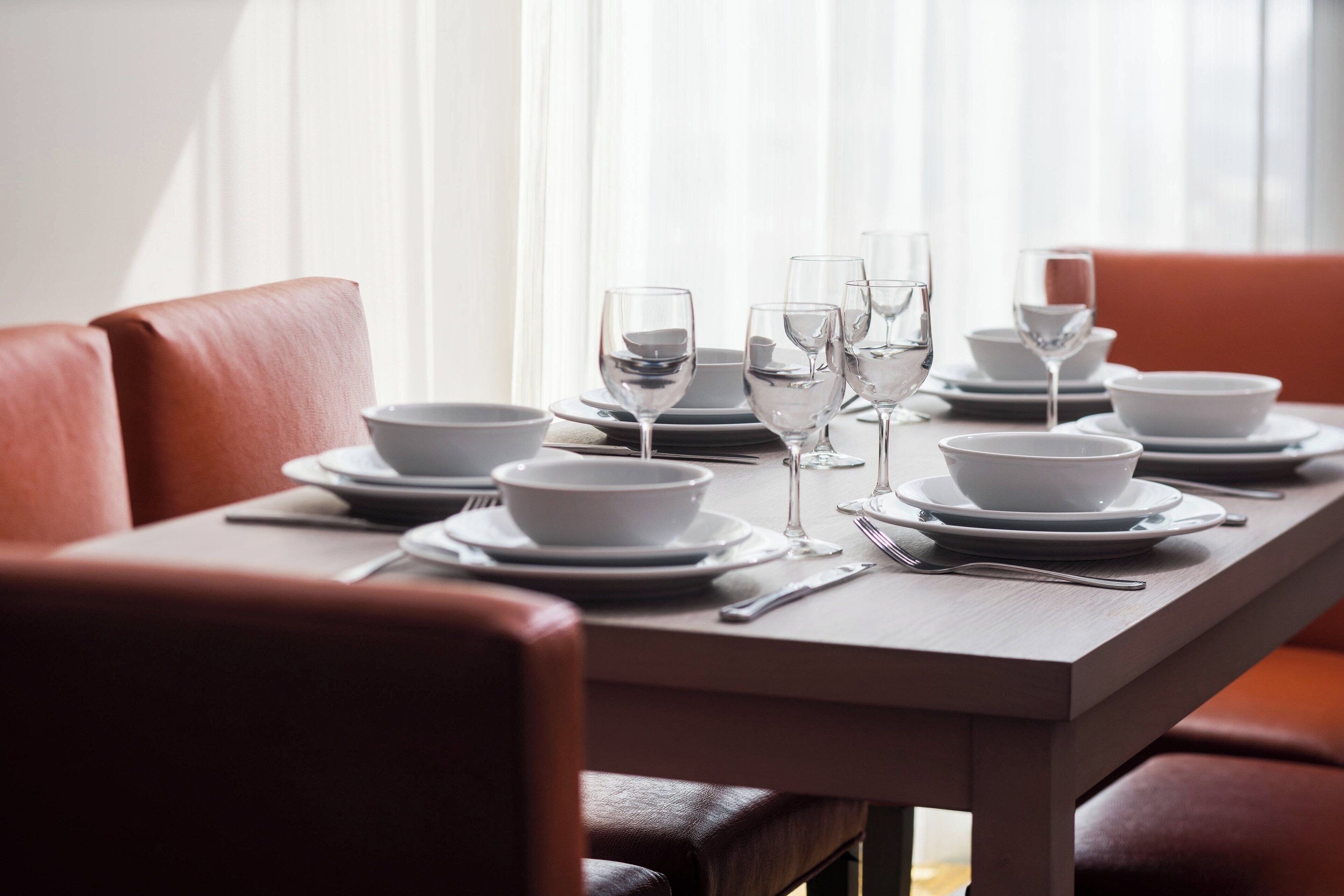 A dining table set with white bowls, plates, and wine glasses, surrounded by red leather chairs.