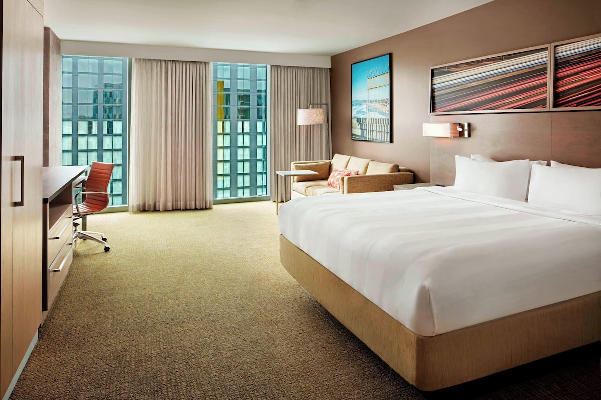 Habitación de hotel con una cama grande, un escritorio, una silla y vistas a un paisaje urbano a través de amplias ventanas.