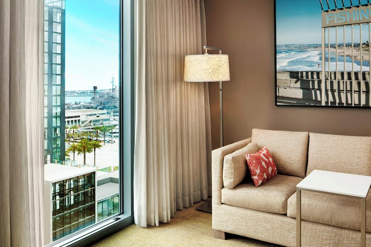 Habitación de hotel con un ventanal amplio que ofrece vistas a la ciudad, un sofá color beige con un cojín rojo, una lámpara de pie y una pequeña mesa blanca.