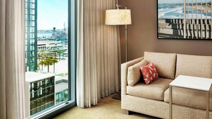 Une chambre d’hôtel dotée d’une grande fenêtre donnant sur la ville, d’un canapé beige agrémenté d’un coussin rouge, d’une lampe sur pied et d’une petite table blanche.