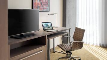 A modern hotel room with a large flat-screen TV, a laptop on a desk, a comfortable office chair, and a large window with sheer curtains.
