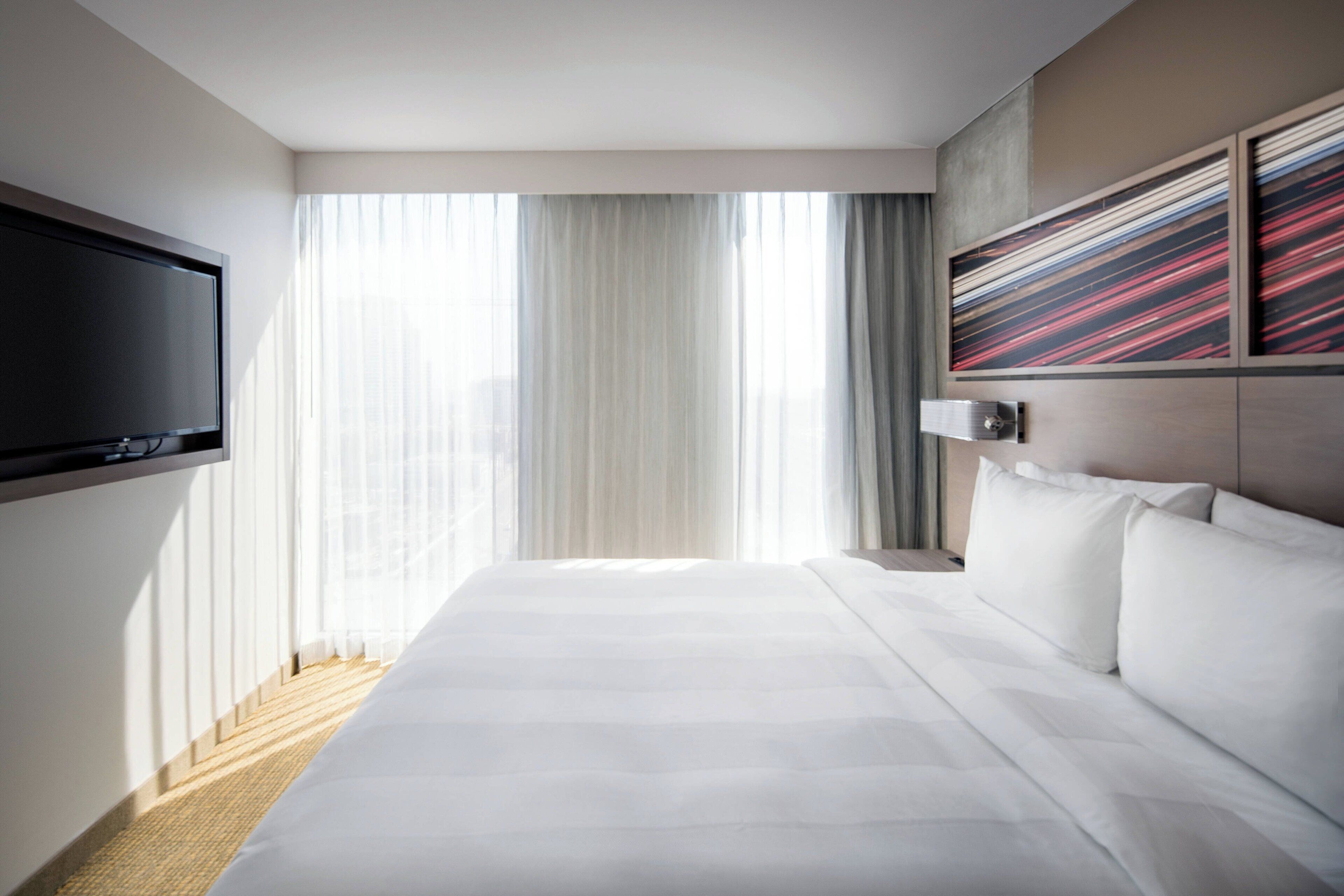 Habitación de hotel con una cama grande, un televisor de pantalla plana en la pared y una ventana con cortinas translúcidas.