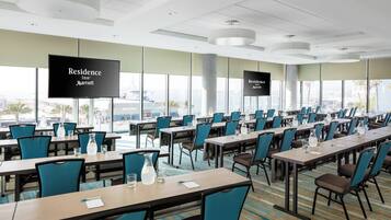 A conference room with multiple tables and chairs, large windows with city views, and screens displaying the hotel name.