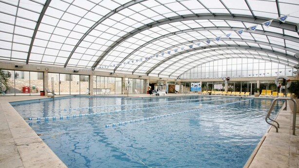 Ein Hallenbad mit Glasdach, deutlich abgetrennten Bahnen und Liegestühlen.