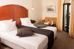 Habitación de hotel con dos camas, cabecera de madera, una mesita con lámpara, un sofá y vistas a través de un ventanal.