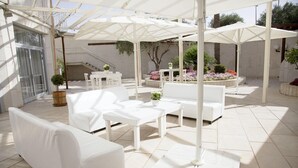 Eine überdachte Terrasse im Freien mit weißen Möbeln, darunter Sofas und Tische, und einer Treppe, die zu einer erhöhten Ebene führt.