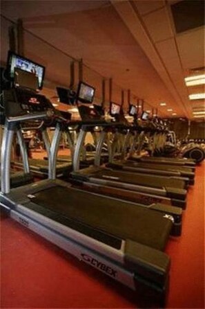 Une rangée de tapis de course dotés d’écrans numériques, dans une salle de sport au sol recouvert d’un tapis rouge.