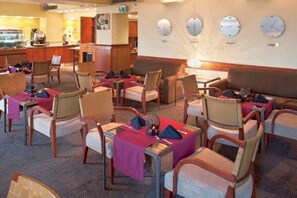 Une salle à manger d’hôtel avec des tables dressées pour les clients, des chaises et une horloge murale.