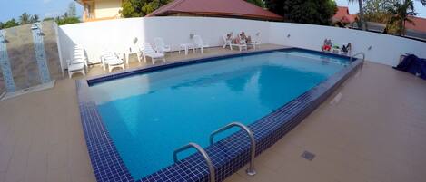 A swimming pool with a tiled edge, surrounded by white lounge chairs and a white wall.