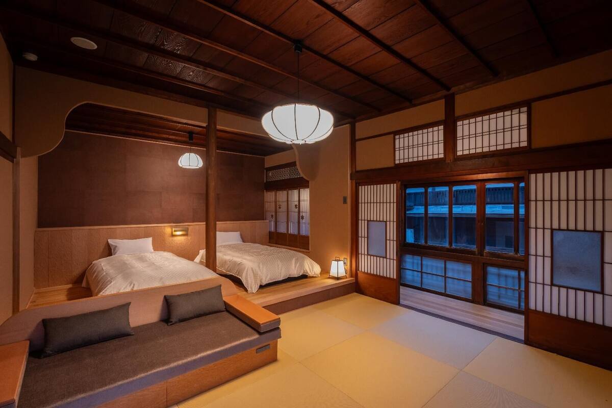 A traditional Japanese-style room with tatami flooring, two beds with white bedding, a low wooden bench, and a hanging light fixture.