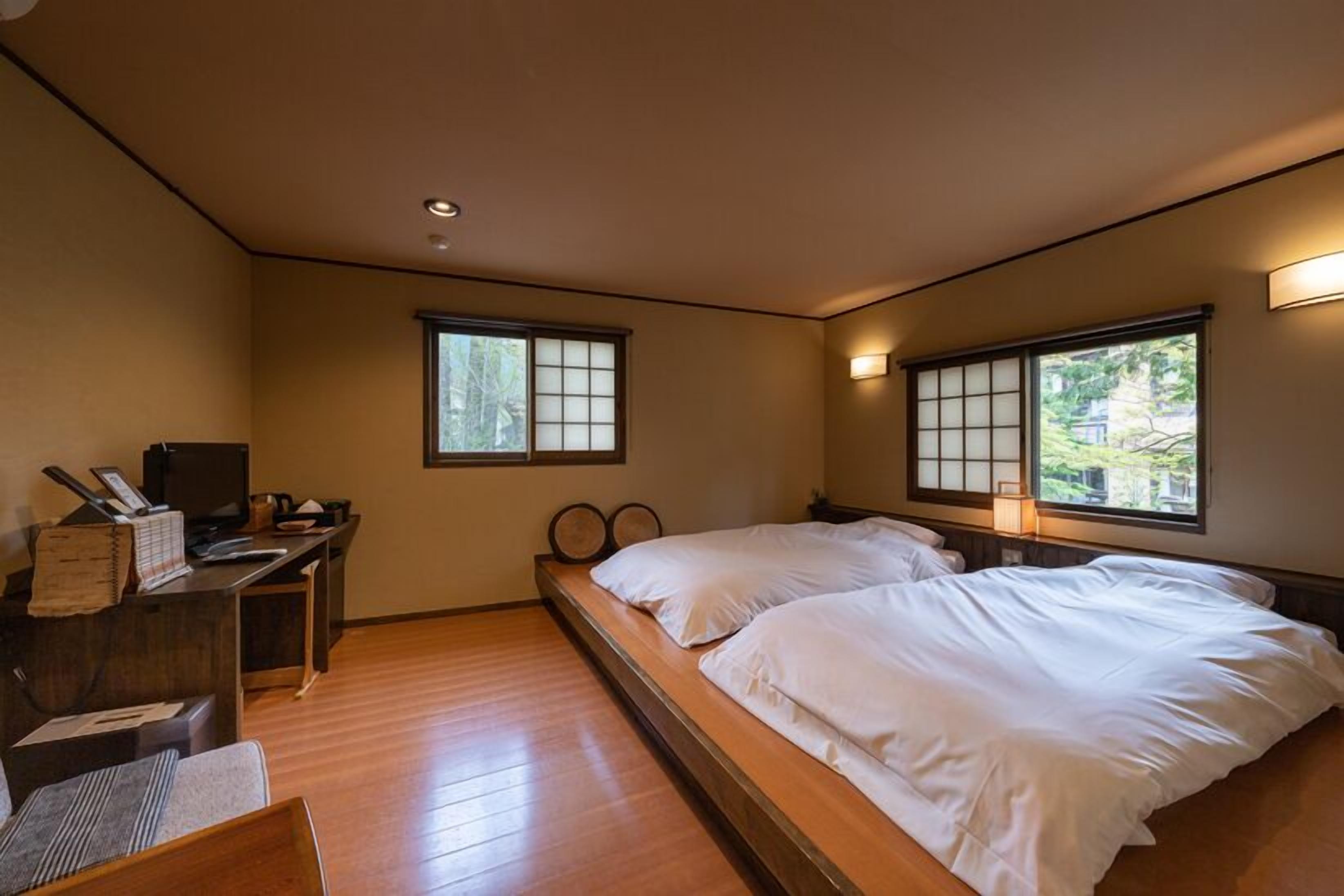 A room with two beds, a desk, a television, and windows with views of greenery.