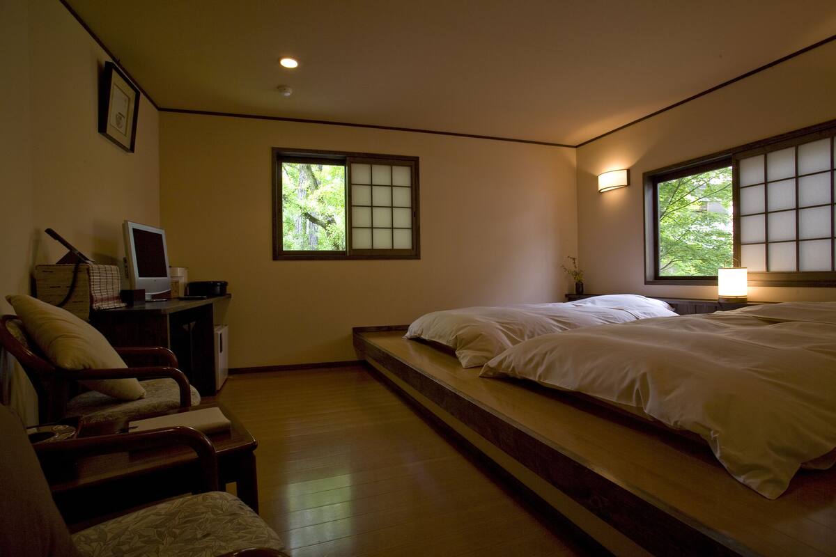 A room with a bed, a chair, a desk with a computer, and a window with a view of trees.