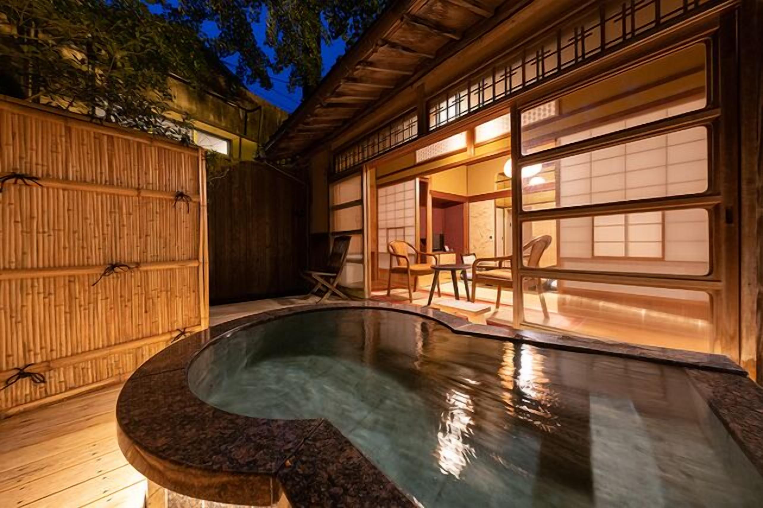A traditional Japanese-style outdoor hot spring area with a wooden deck, a stone hot tub, and a view of a building with sliding doors.