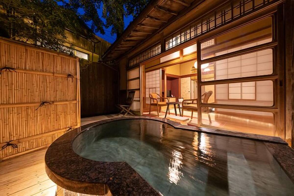 A traditional Japanese-style outdoor hot spring area with a wooden deck, a stone hot tub, and a view of a building with sliding doors.