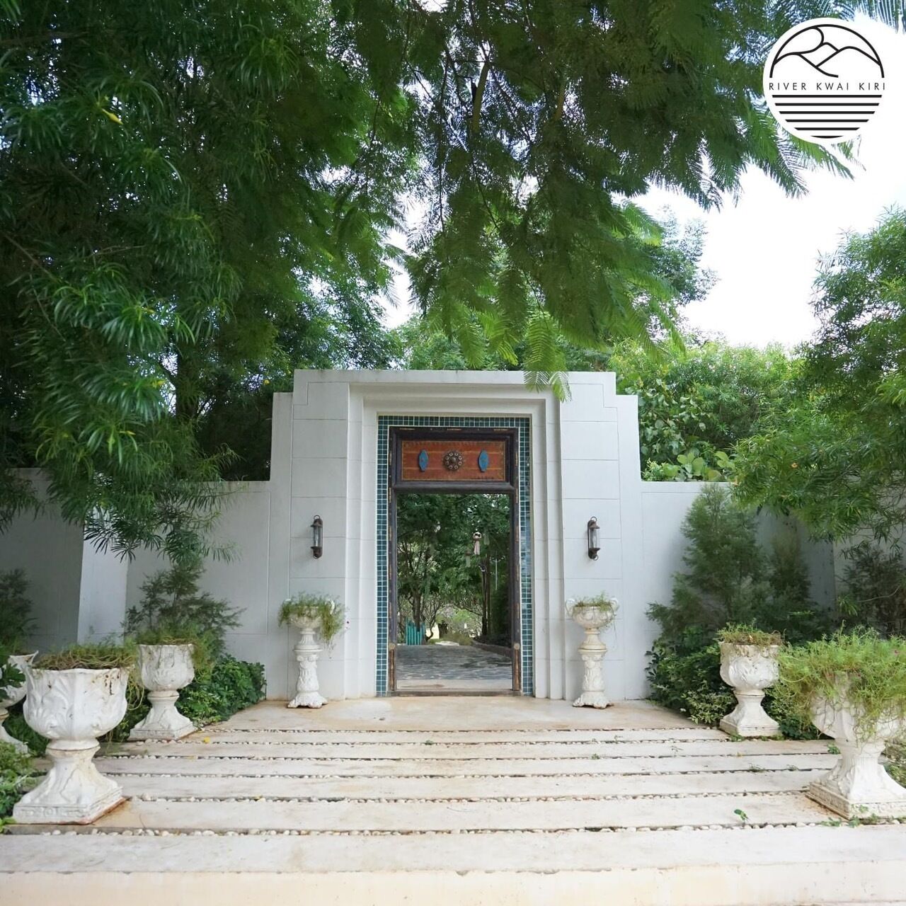 Una puerta blanca con arco decorativo que da a un sendero rodeado de abundante vegetación.