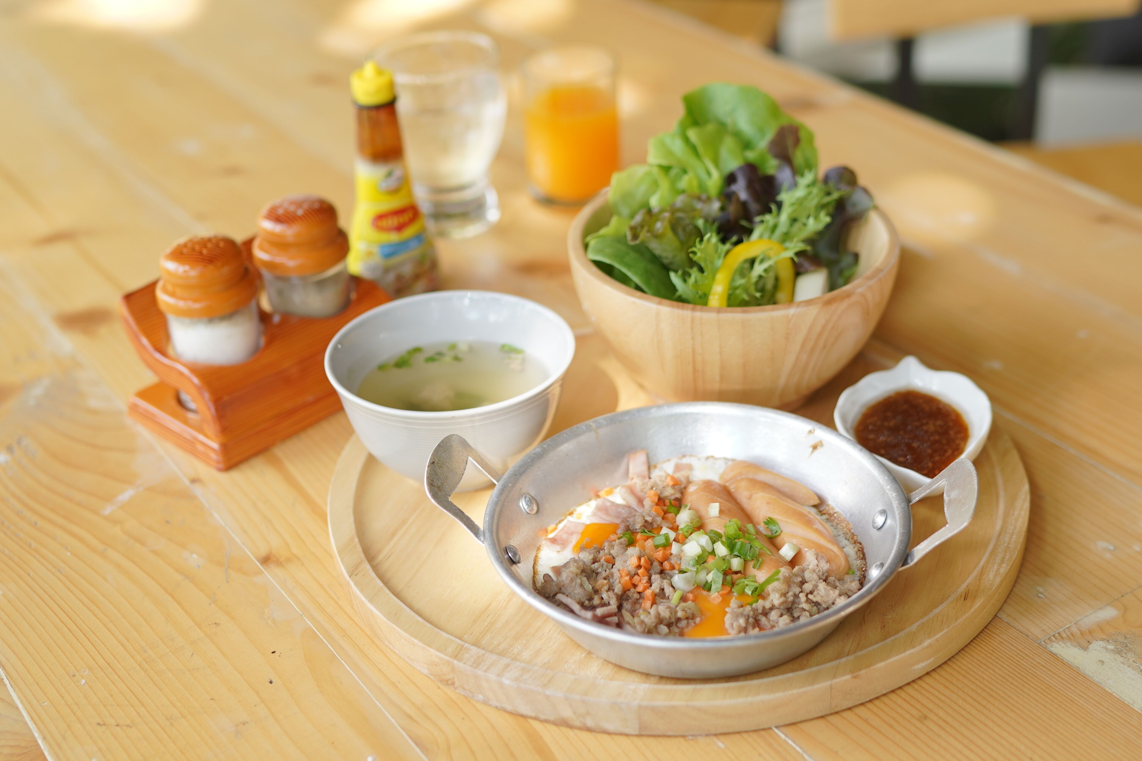 A meal with a bowl of salad, a bowl of soup, a pan with meat and vegetables, condiments, and a bottle of sauce on a wooden table.