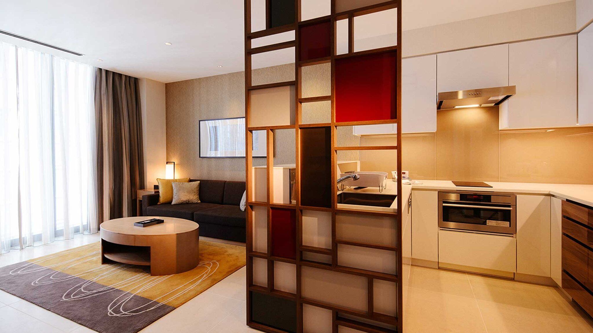 Una habitación de hotel moderna con sala de estar, sofá, mesa de centro, cocina con estufa y horno, y un ventanal con cortinas.
