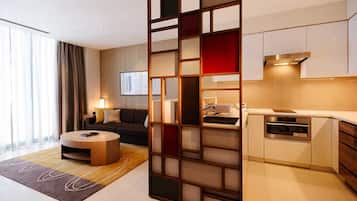 A modern hotel room with a living area, a sofa, a coffee table, a kitchen with a stove and oven, and a large window with curtains.