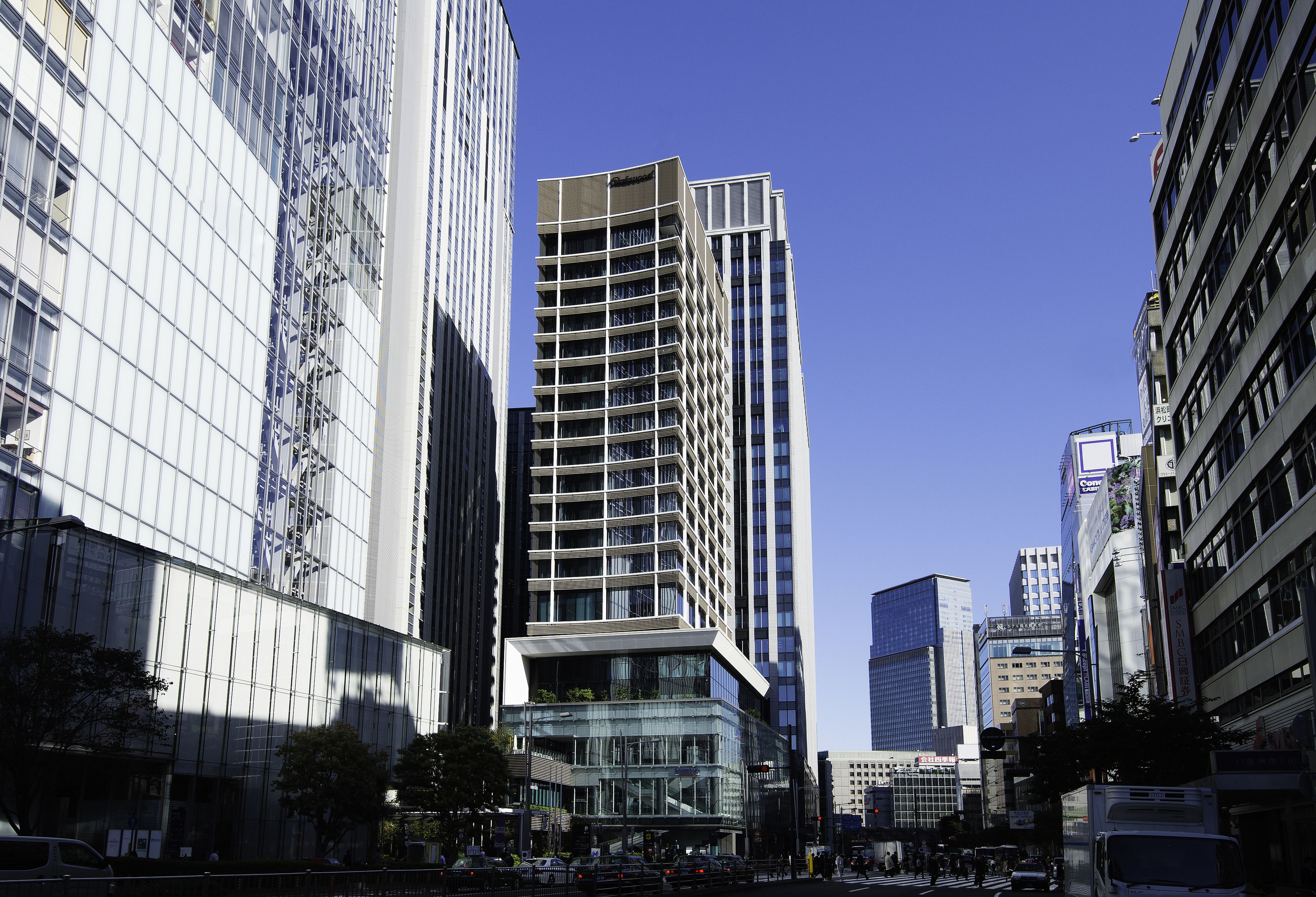 A modern urban street with tall buildings, including a prominent glass-fronted structure and a high-rise with a unique stepped design.