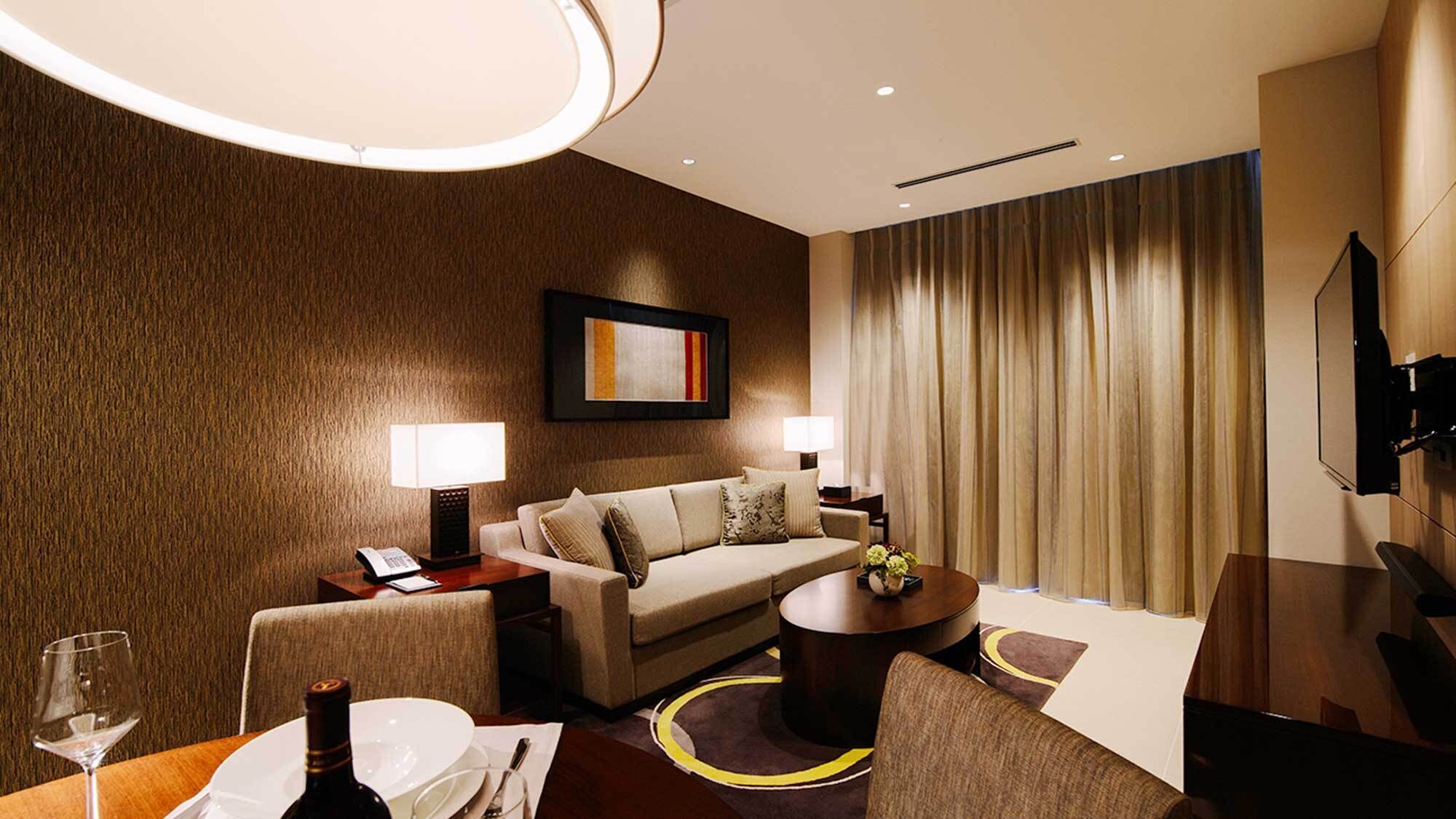 A hotel room with a sofa, two bedside tables with lamps, a coffee table, a dining table with a wine glass and bottle, and a TV mounted on the wall.