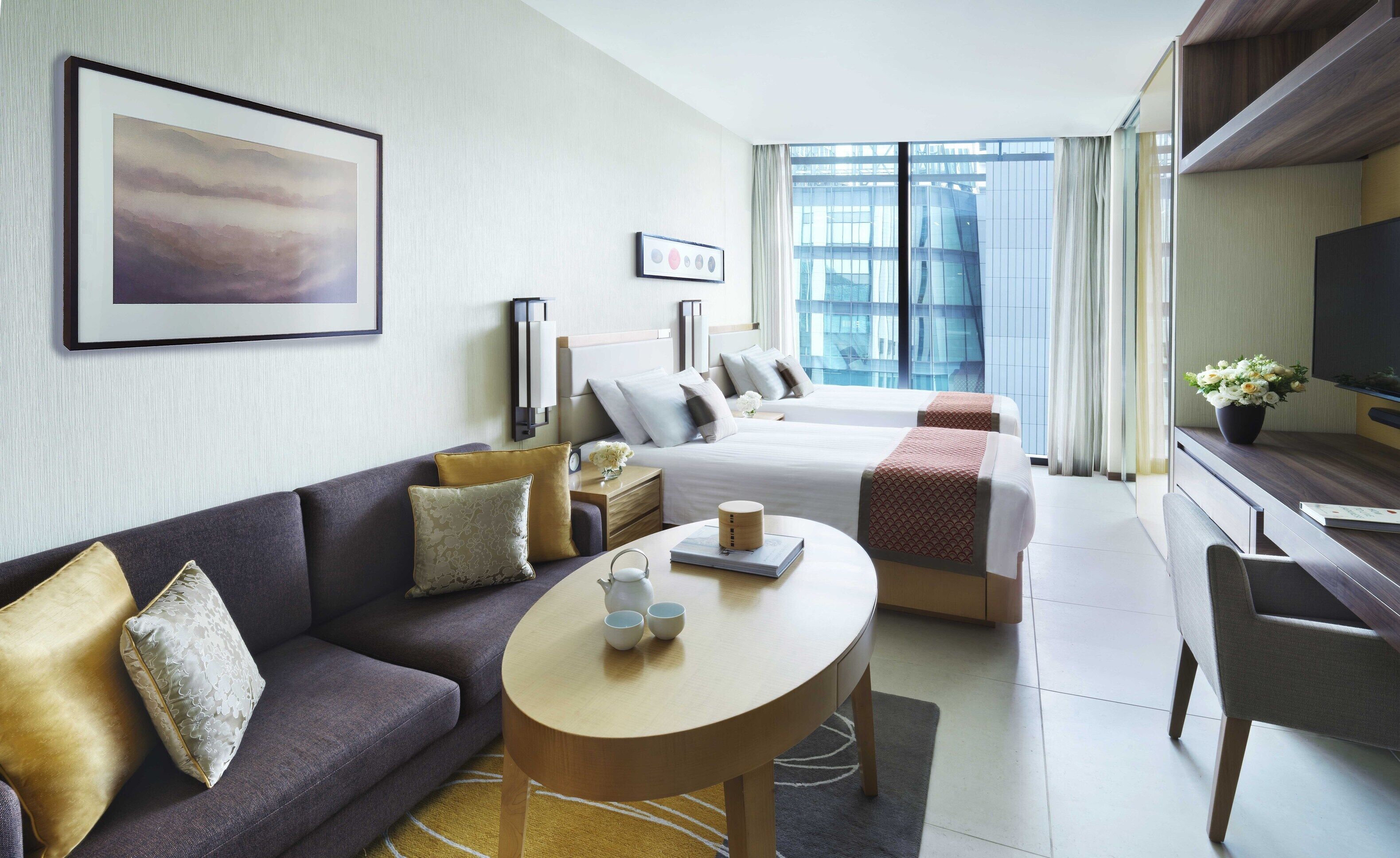 A hotel room with a sofa, two beds, a TV, a desk, and a large window with a city view.