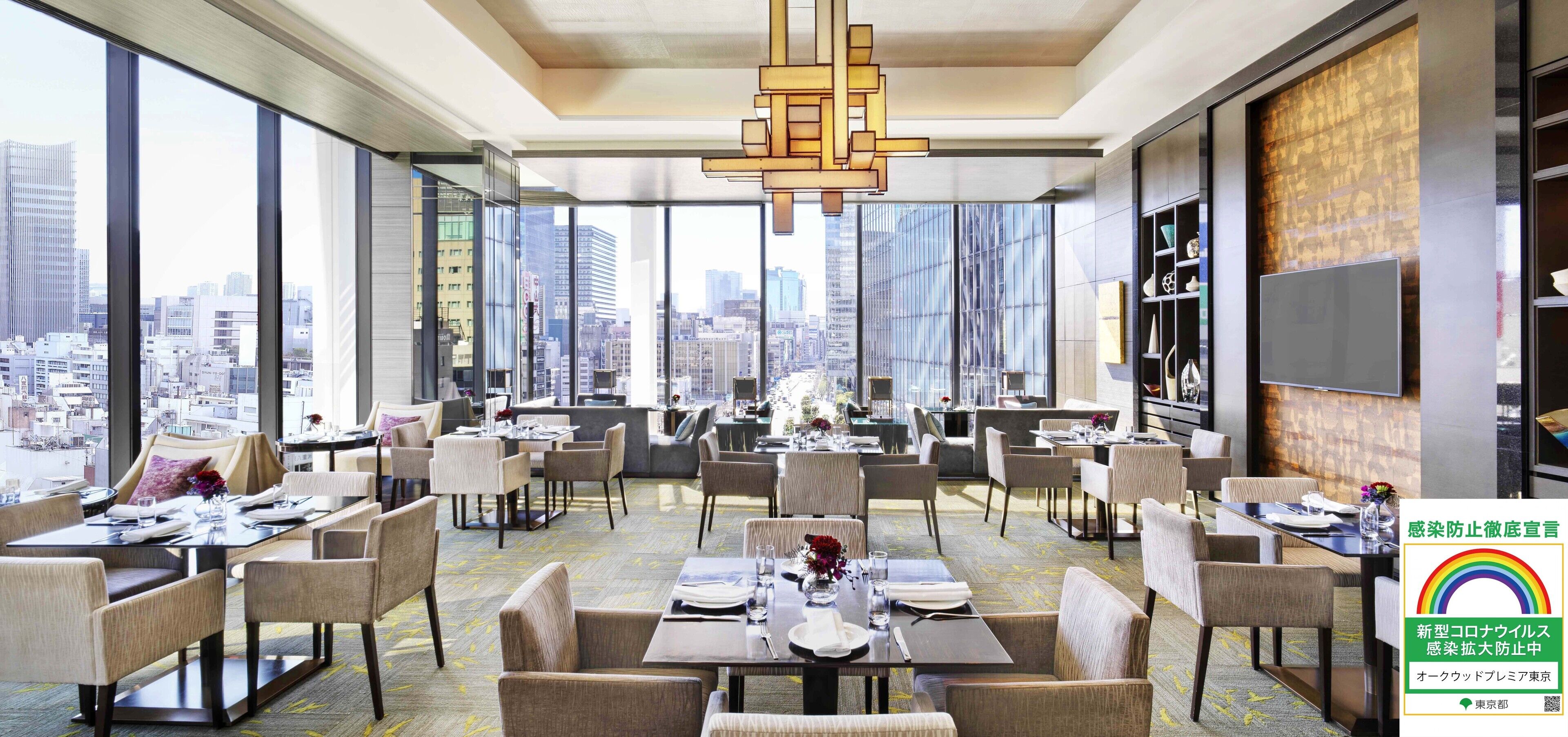 Un comedor moderno con mesas dispuestas para los huéspedes, amplias ventanas con vista a la ciudad y una lámpara decorativa.