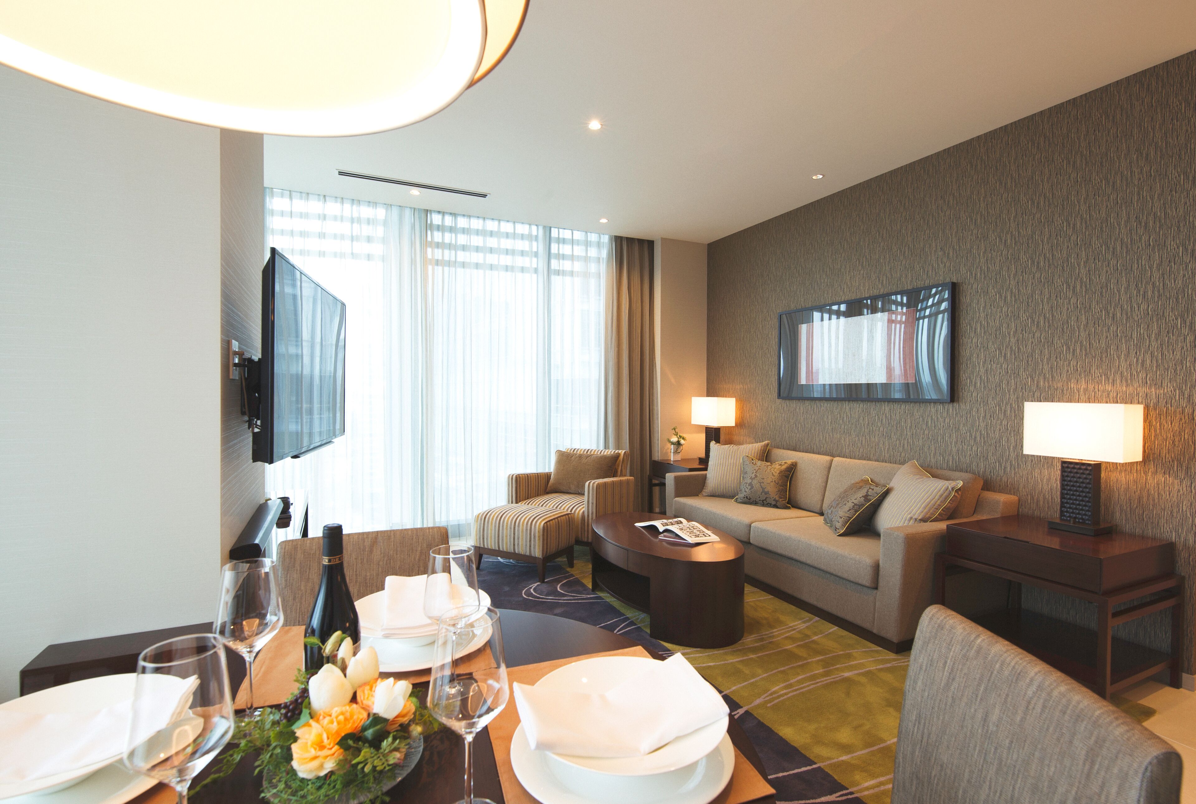 A modern hotel room with a dining area, a sofa, a TV, and a large window with curtains.