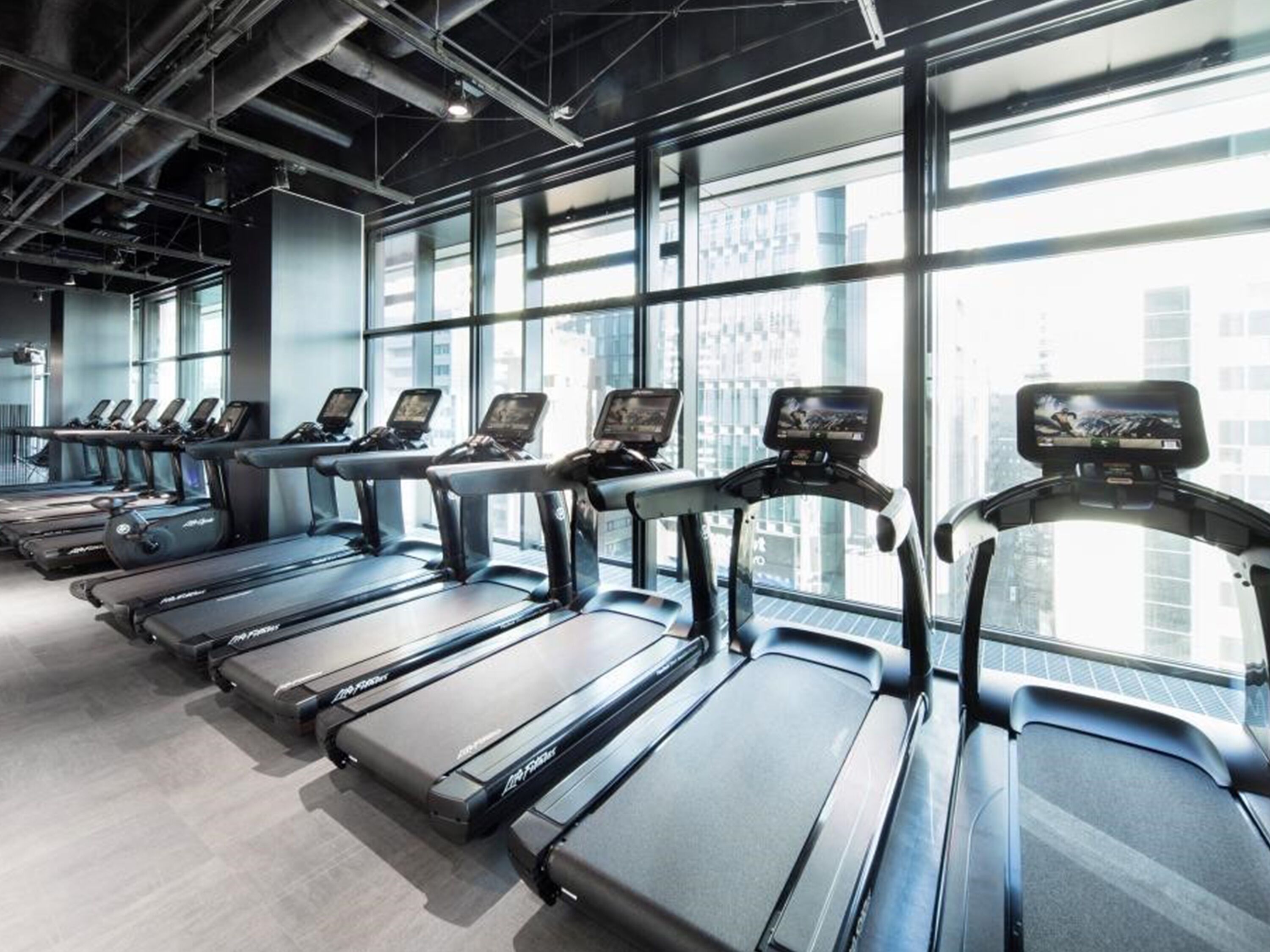 Une rangée de tapis de course dans une salle de sport dotée de grandes fenêtres et offrant une vue sur la ville.