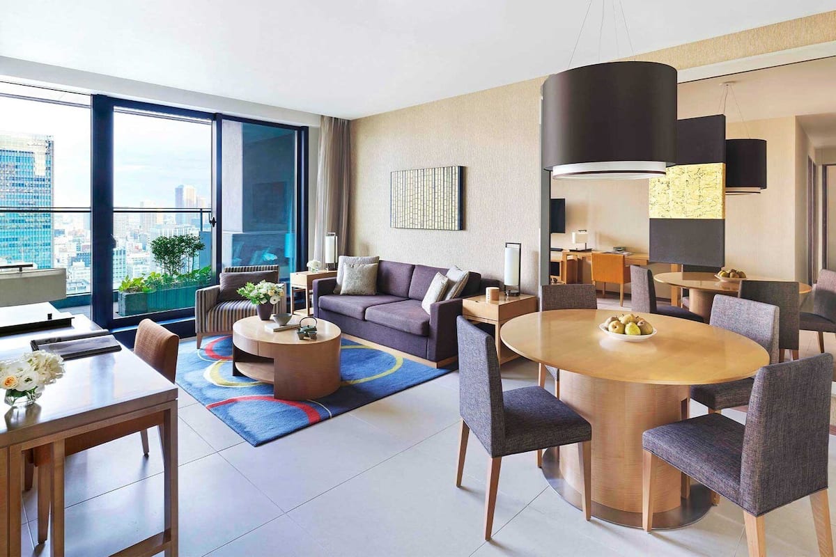 Habitació d'hotel moderna amb zona de saló, menjador i una finestra gran amb vista a la ciutat.