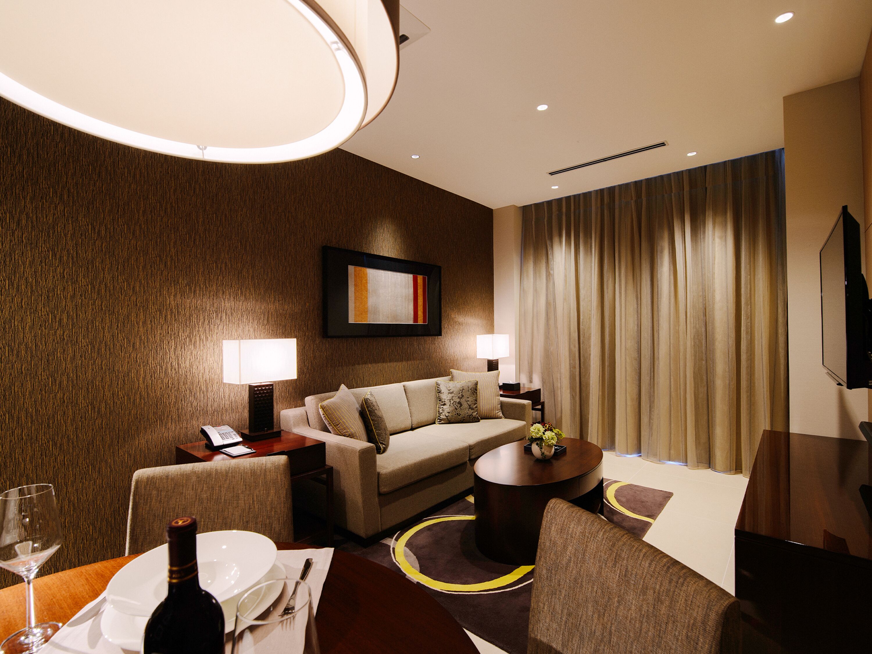 A hotel room with a sofa, a dining table set with a bottle and glasses, a coffee table, and a TV.