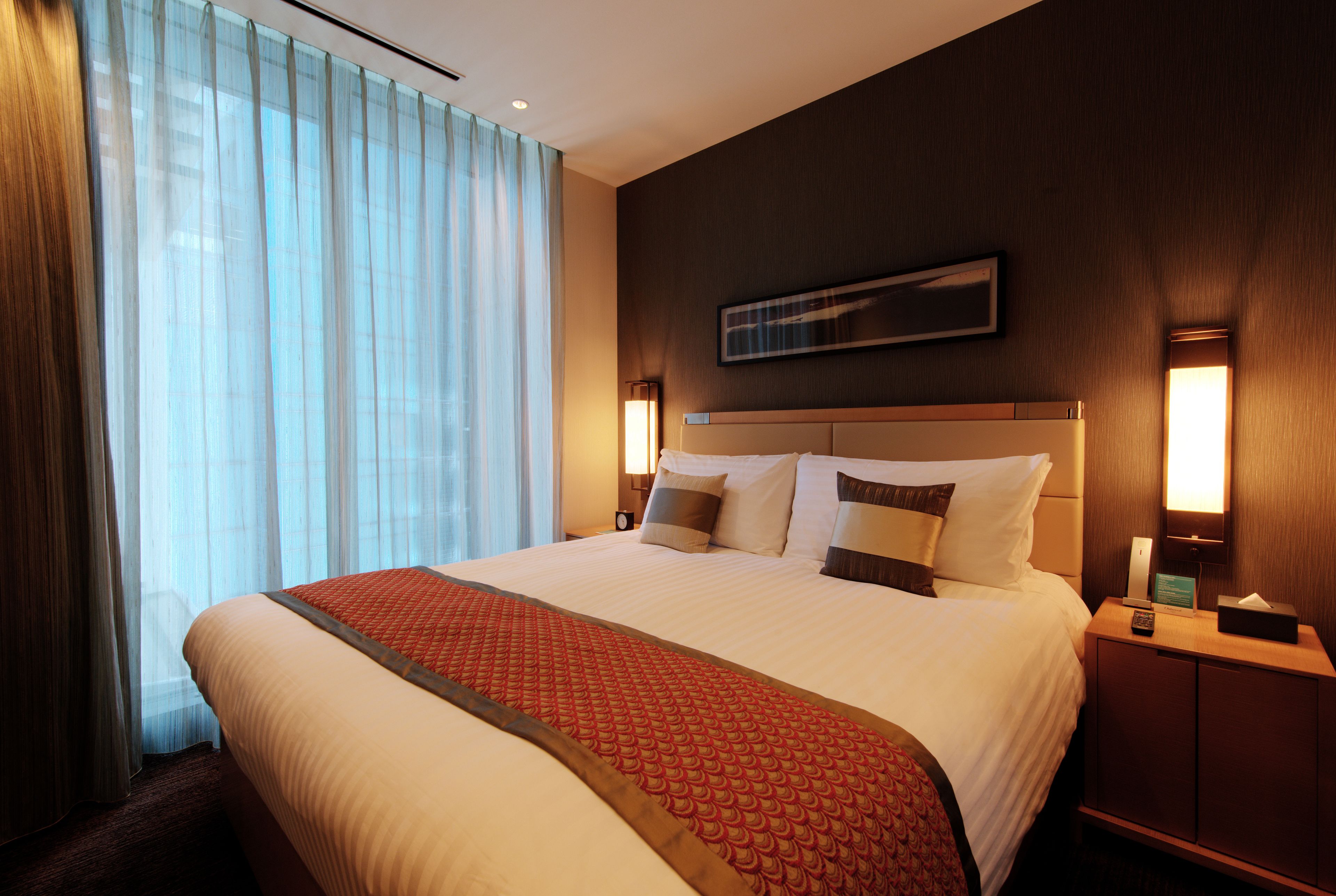 A hotel room with a large bed, a headboard with a flat panel, two bedside tables with lamps, and a window with sheer curtains.