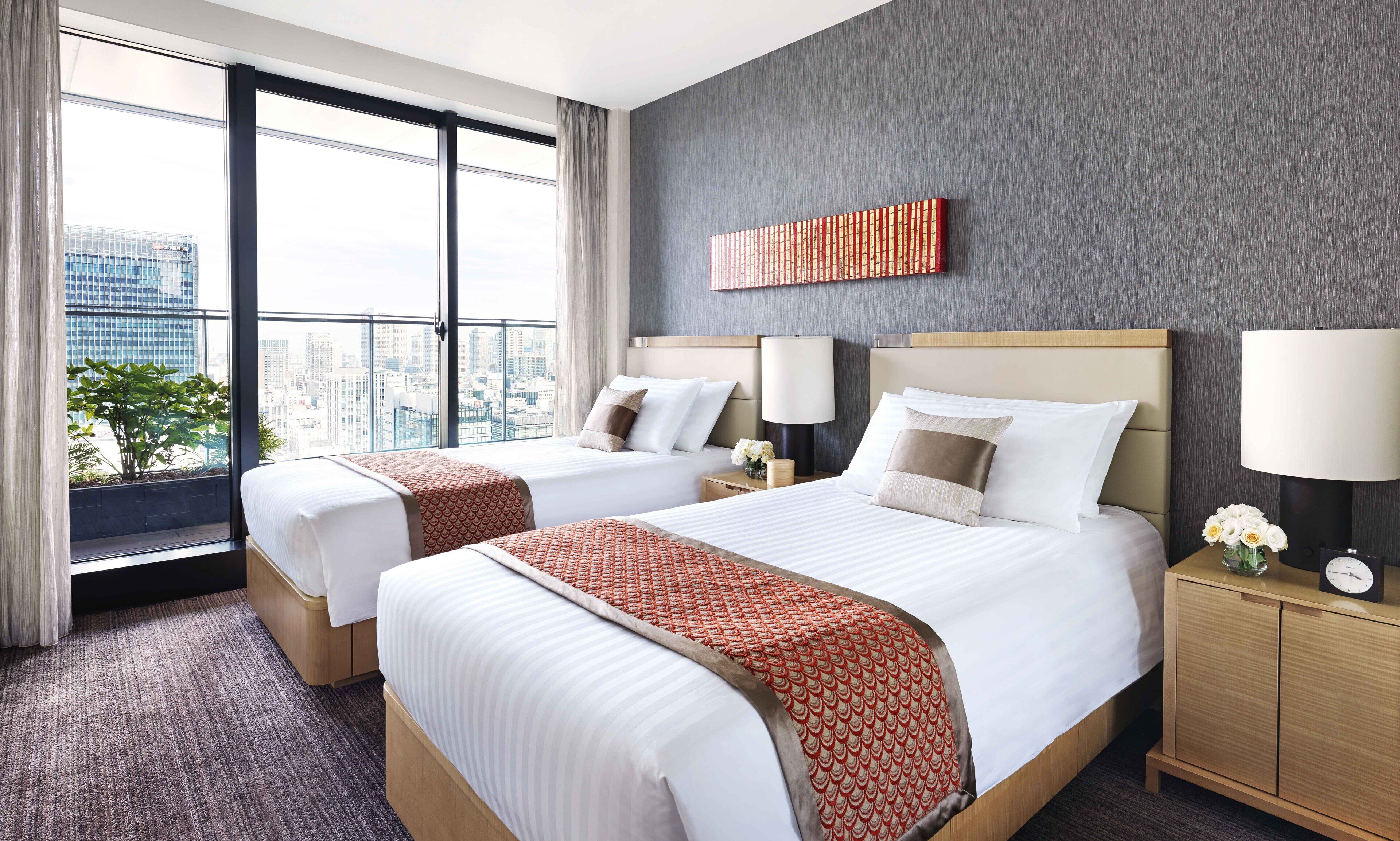 A hotel room with two beds, a large window with a city view, a grey textured wall, and a modern design.